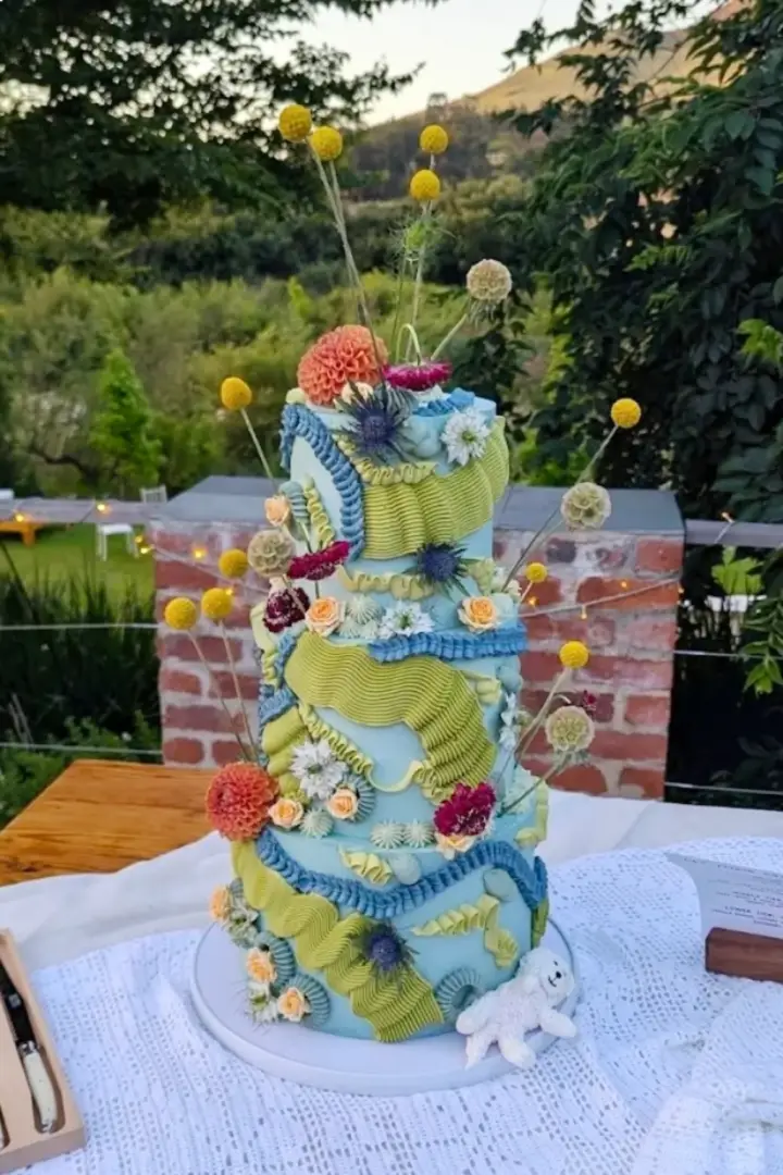 A vibrant, multi-tiered cake with blue icing is adorned with intricate green and blue ruffles and colorful flowers - by Crumb Boutique Bakery, Cape Town